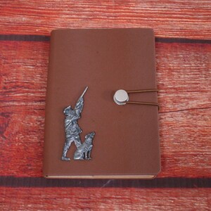 May include: Brown leather journal with a silver metal design of a hunter and dog. The journal has a silver metal closure with a brown leather strap.