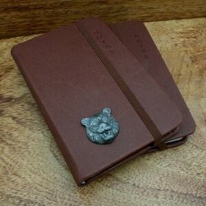 Puede incluir: Dos cuadernos de cuero marrón con la palabra "NOTES" grabada en el lomo. Cada cuaderno tiene una banda elástica marrón y un emblema de cabeza de tigre plateado. Los cuadernos están sobre una superficie de madera.