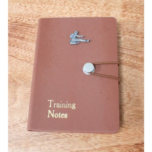 Op de afbeelding: Een bruin lederen gebonden dagboek met een zilveren vechtsportfiguur op de voorzijde. Het dagboek heeft een gouden inscriptie met de tekst "Training Notes".