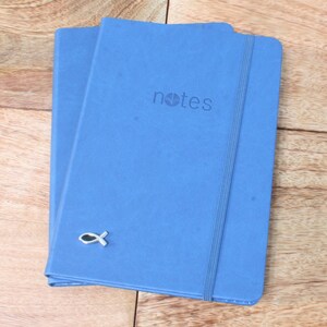 May include: Two blue notebooks with a silver fish charm on the front. The top notebook has the word "notes" printed on the front.