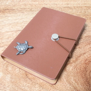 May include: A brown leather journal with a silver turtle charm and a silver button closure. The journal has a brown elastic band closure.