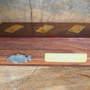 Puede incluir: Una caja de madera con una placa de latón que dice "Su mensaje grabado aquí gratis". La caja tiene un diseño de pez plateado en la parte delantera y tres dominós incrustados en la parte superior.