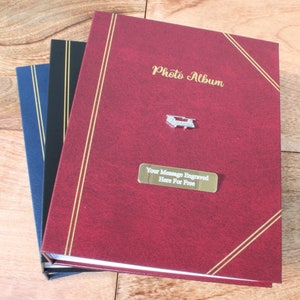 Könnte beinhalten: Eine Auswahl an Fotoalben in Rot, Schwarz und Blau. Das rote Album trägt den goldenen Schriftzug "Photo Album", ein silbernes Emblem und eine goldene Plakette mit der Aufschrift "Your Message Engraved Here For Free".