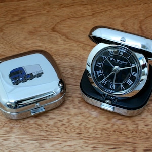 May include: A silver travel alarm clock with a black face and Roman numeral hour markers. The clock is in a silver case with a blue truck design on the lid. The clock face reads "WM. WIDDOP".