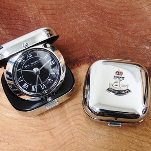 Könnte beinhalten: Eine silberne Taschenuhr mit schwarzem Zifferblatt und römischen Ziffern. Die Uhr befindet sich in einem silbernen Etui mit einem Royal Navy Emblem auf dem Deckel. Die Uhr zeigt "WM. WIDDOP" und "EST. 1887".