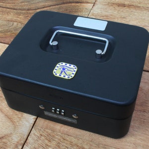 May include: Black metal cash box with a combination lock and a handle. The box has a gold sticker with a blue eye of Horus symbol.