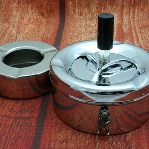 May include: Two chrome ashtrays on a wooden surface. One is a simple, open bowl. The other is a covered ashtray with a black handle and a small astronaut figure on the side. The ashtrays are shiny and reflective.