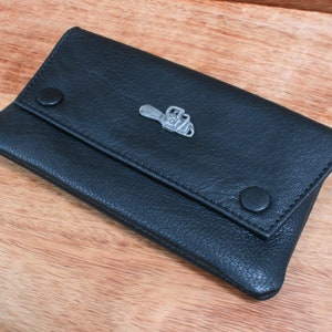 May include: A black leather tobacco pouch with a silver metal design of a chainsaw on the front. The pouch has a flap that closes with two snaps.