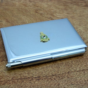 Peut inclure: Un porte-cartes en métal argenté avec un emblème or et vert. L'emblème présente une couronne et un cercle avec une couronne de laurier verte et une étoile dorée.