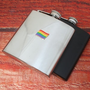 May include: Two stainless steel hip flasks, one with a rainbow flag design. The flasks are on a wooden surface.