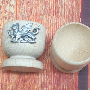 May include: Two wooden egg cups. One cup features a silver-colored dragon emblem. The cups are made of light-colored wood and have a rounded shape. The egg cups are displayed on a wooden surface.