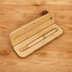 K&ouml;nnte beinhalten: Ein helles Holzstift- und Etui-Set. Der Stift hat eine goldfarbene Spitze und Akzente. Der Stift befindet sich in einem hellen Holzgeh&auml;use mit einem Klappdeckel. Das Geh&auml;use ist ge&ouml;ffnet und zeigt den Stift.