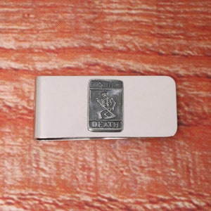 以下が含まれることがあります： 死神のタロットカードの白黒画像が刻まれたシルバーのマネークリップ。カードには、鎌を持った骸骨の姿が描かれており、画像の下には「DEATH」という文字が印刷されています。