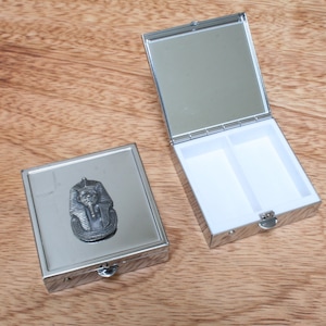 Könnte beinhalten: Eine silberne Pillendose mit einem Scharnierdeckel und zwei Fächern. Die Vorderseite der Dose zeigt ein erhabenes Bild eines ägyptischen Pharaos.