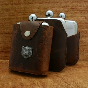 May include: Three silver hip flasks with leather sheaths. The flask on the left has a bear head design on the front.