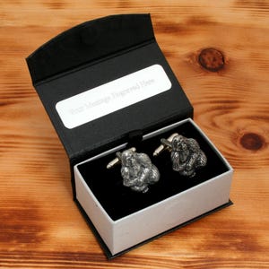 Puede incluir: Una caja de regalo negra con una inscripción plateada que dice "Su mensaje grabado aquí". La caja contiene dos gemelos de plata con un diseño detallado de un orangután.