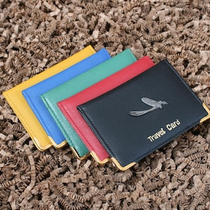 Op de afbeelding: Een stapel kleurrijke reispashouders in geel, blauw, groen, rood en zwart. De zwarte houder heeft een zilveren vogel-embleem en de woorden "Travel Card" in goud. Elke houder heeft goudkleurige hoekaccenten.