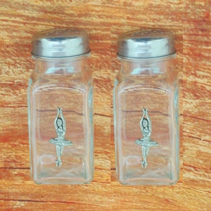 Könnte beinhalten: Zwei klare Glassalz- und Pfefferstreuer mit silbernen Deckeln. Jeder Streuer zeigt eine Ballerina-Figur aus Zinn.