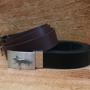 Op de afbeelding: Twee leren riemen, één opgerold en één plat, met een zilveren gesp met een rennende vos. De opgerolde riem is donkerbruin en de platte riem is zwart. De riemen worden gepresenteerd op een houten oppervlak.