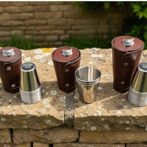 Pode incluir: Um conjunto de copos de dados de couro castanho com detalhes em metal prateado e um emblema de urso na tampa. O conjunto inclui copos de dados de aço inoxidável. O número 10 é impresso nos copos.