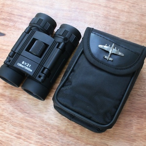 May include: Black binoculars with a silver airplane design on the case. The binoculars have a 8x21 magnification and a 128m/1000m field of view.