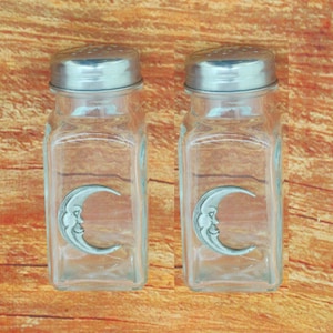 May include: A set of two glass salt and pepper shakers with pewter crescent moon designs. The shakers are clear glass with silver lids.