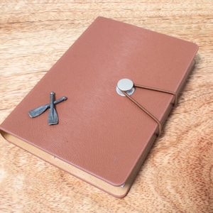May include: A brown leather-bound journal with a silver clasp and two crossed oar-shaped metal accents. The journal has a textured surface and a brown elastic band closure.