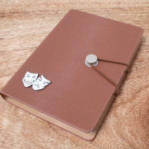 May include: A brown leather-bound journal with a silver button closure and a decorative elastic band. The journal has two silver metal masks on the front, one smiling and one frowning.