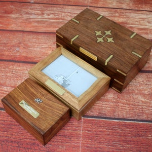 Op de afbeelding: Drie houten dozen met messing accenten. De grootste doos heeft een decoratief messing ontwerp op de deksel. De middelste doos is een fotolijst met een zwart-wit foto van een zeilboot. De kleinste doos heeft een messing plaatje met de tekst "60 jaar aan herinneringen 1960 tot 2020".