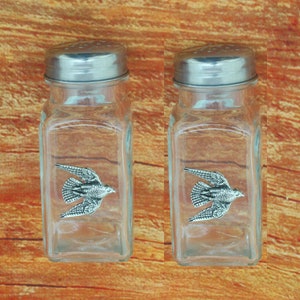 May include: Two clear glass salt and pepper shakers with silver lids. Each shaker features a pewter bird design.