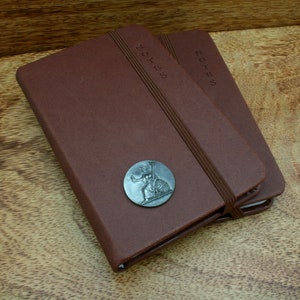 May include: Two brown leather notebooks with the word "NOTES" embossed on the cover. Each notebook has an elastic band closure and a decorative metal emblem. The notebooks are on a wooden surface.