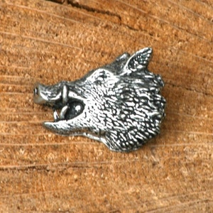 May include: A silver-colored pewter pin depicting the head of a wild boar. The boar's head is facing to the left, with its mouth open and teeth visible. The pin is detailed with textured fur and a realistic depiction of the boar's features.
