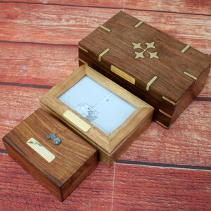 May include: Three wooden boxes with brass accents. The largest box has a decorative brass design on the top. The middle box has a glass window and a brass plaque. The smallest box has a brass plaque and a small metal tractor on top.