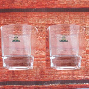 May include: Two clear glass tumblers with a gold and green logo on the front. The tumblers are sitting on a wooden surface.