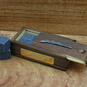 May include: A wooden box with a hinged lid, containing a set of grey whisky stones and a silver-coloured rifle figurine. The box has a gold plaque with the text "Your Message Engraved Here For You Free".