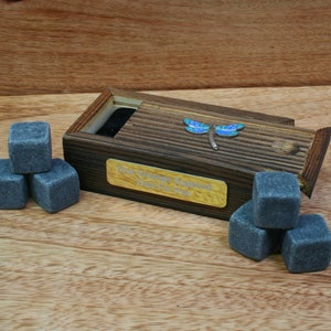 Puede incluir: Una caja de madera con tapa deslizante, con una incrustación de libélula y una placa de latón con la inscripción "To William Reginald Have a Great Time". La caja contiene un juego de piedras de whisky grises.