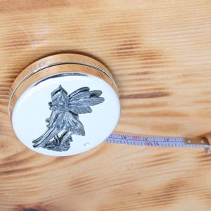 May include: A silver and white measuring tape with a fairy design on the top. The tape measure is extended, showing inch markings. The background is a light brown wooden surface.