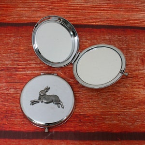 May include: A silver compact mirror with a hare design on the front. The mirror is open and the inside is reflective.