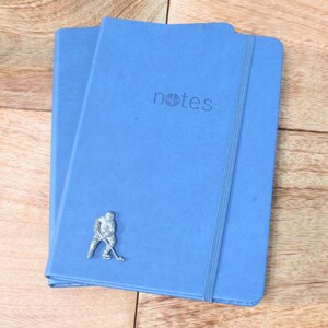 May include: Two blue notebooks with a silver hockey player figurine on the front. The notebook on top has the word "notes" printed on the front.