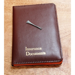 May include: A brown leather folder with the words "Insurance Documents" embossed in gold lettering. A silver whistle pin is attached to the front of the folder.