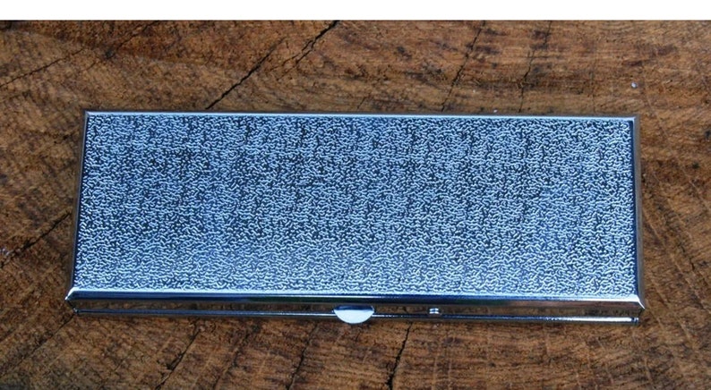 May include: A silver metal rectangular box with a textured surface. The box has a hinged lid and a small latch.