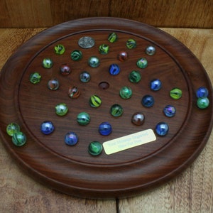 Könnte beinhalten: Ein Holzspielbrett mit einer runden Spielfläche und einem zentralen Loch. Das Brett ist von einem erhöhten Rand umgeben und hat eine kleine Plakette mit dem Text "Deine Erinnerungen verewigt, ein Schuss nach dem anderen". Das Brett ist mit bunten Murmeln in Grün-, Blau- und Rottönen gefüllt.