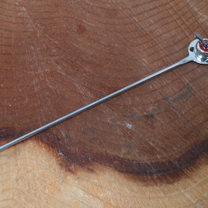 May include: A silver metal stirring stick with a round ball on one end and a propeller-shaped top on the other end. The propeller is red and white.