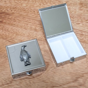 Könnte beinhalten: Eine silberne Pillendose mit einem Pinguin-Design auf dem Deckel. Die Dose hat zwei Fächer im Inneren.