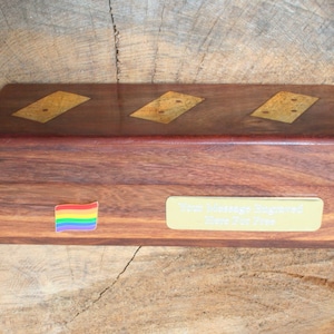 May include: A wooden box with a rainbow flag sticker and a gold plaque that reads "Your Message Engraved Here For Free". The box has three inlaid wooden squares with a pattern of dots.