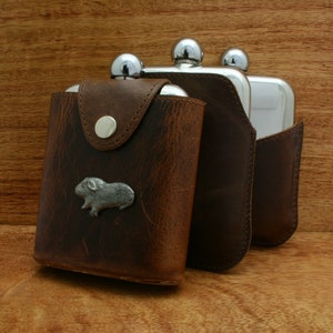 May include: Three stainless steel hip flasks with brown leather pouches. The flask on the left has a small silver guinea pig charm.