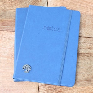May include: Two blue notebooks with a silver tree charm on the front. The top notebook has the word "notes" printed on the front.
