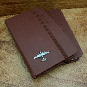 Könnte beinhalten: Zwei braune, ledergebundene Notizbücher mit Gummibändern. Das obere Notizbuch hat einen silbernen Flugzeug-Anhänger an der Vorderseite. Die Notizbücher sind auf den Vorderseiten mit "NOTES" beschriftet.