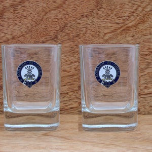 May include: Two clear glass shot glasses with a blue and gold crest on the front. The crest features a white background with a yellow and gold design and the text "Aonaidh Ri Cheile".