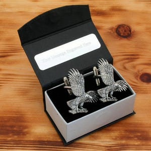 Puede incluir: Un par de gemelos de plata en una caja de regalo negra y plateada. Los gemelos presentan un diseño detallado de un halcón con las alas extendidas. La caja tiene una placa de plata con el texto "Su mensaje grabado aquí".